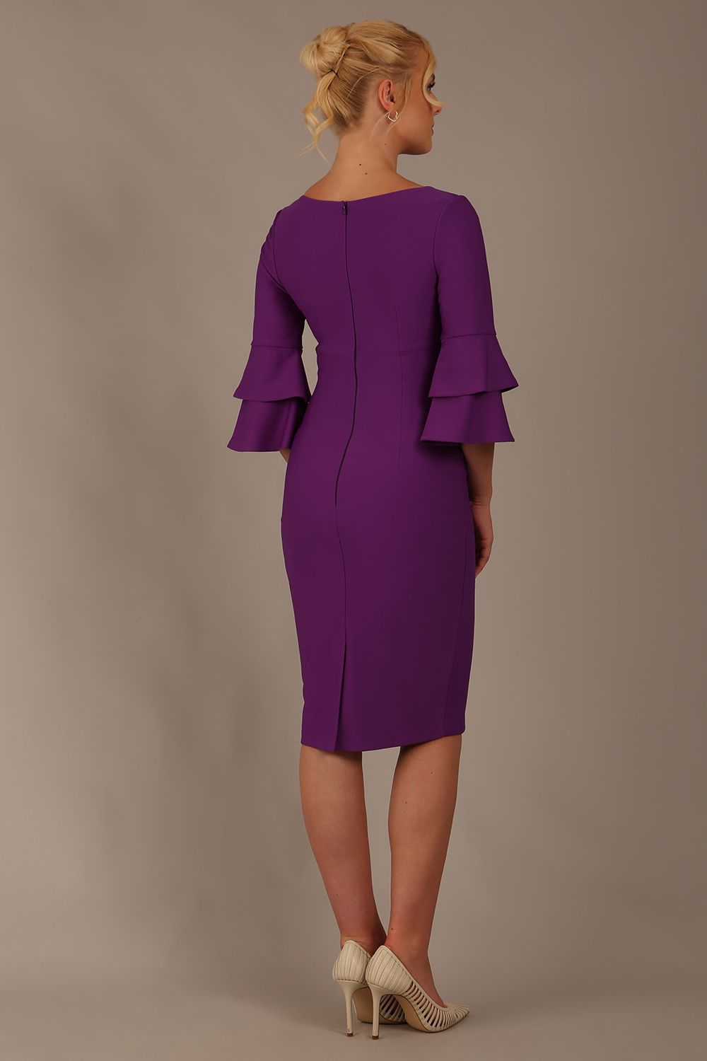A woman wearing a royal purple knee-length diva catwalk meridian pencil dress with a round neckline, double layered peplum three quarter length sleeves, and standing against a plain background.