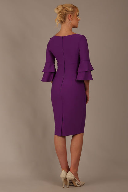 A woman wearing a royal purple knee-length diva catwalk meridian pencil dress with a round neckline, double layered peplum three quarter length sleeves, and standing against a plain background.