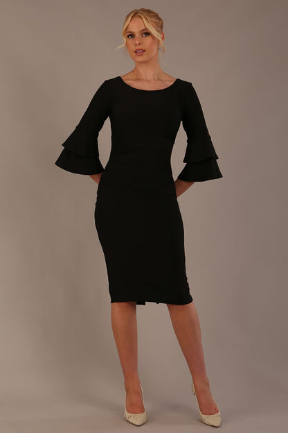 A woman wearing a black knee-length diva catwalk meridian pencil dress with a round neckline, double layered peplum three quarter length sleeves, and standing against a plain background.