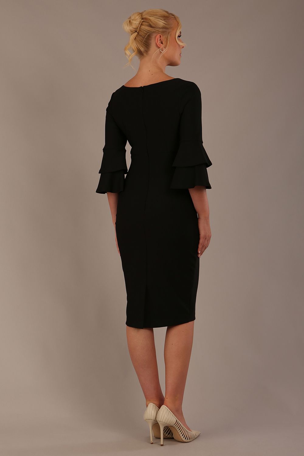 A woman wearing a black knee-length diva catwalk meridian pencil dress with a round neckline, double layered peplum three quarter length sleeves, and standing against a plain background.