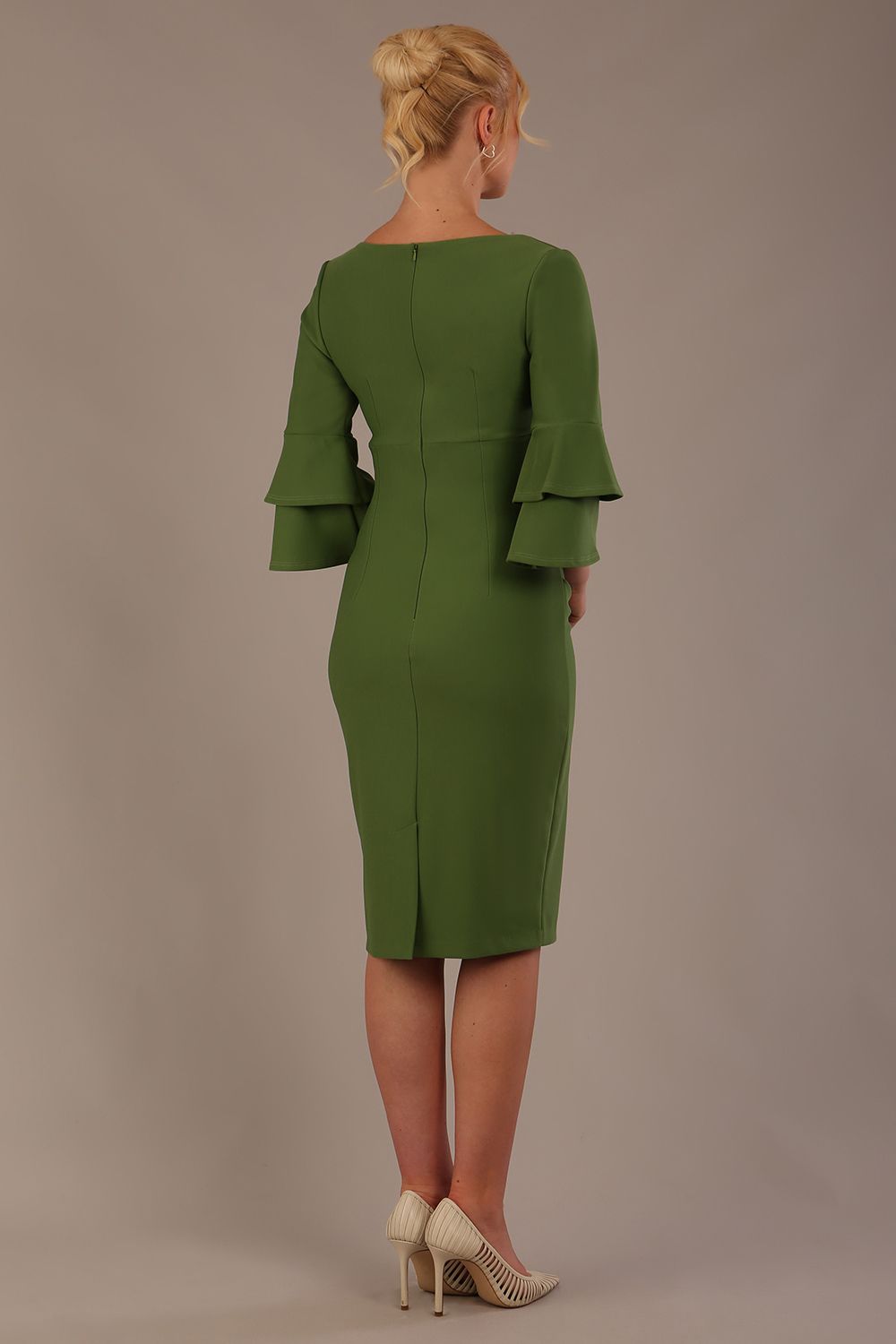 A woman wearing a vineyard green knee-length diva catwalk meridian pencil dress with a round neckline, double layered peplum three quarter length sleeves, and standing against a plain background.