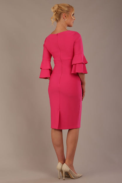 A woman wearing a hibiscus pink knee-length diva catwalk meridian pencil dress with a round neckline, double layered peplum three quarter length sleeves, and standing against a plain background back view