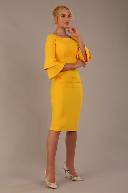 A woman wearing a sunshine yellow knee-length diva catwalk meridian pencil dress with a round neckline, double layered peplum three quarter length sleeves, and standing against a plain background.