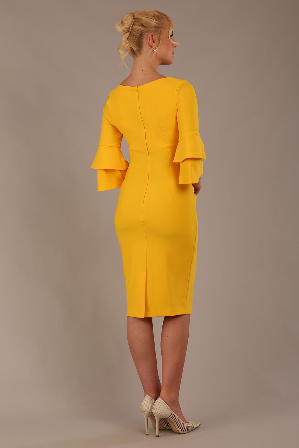 A woman wearing a sunshine yellow knee-length diva catwalk meridian pencil dress with a round neckline, double layered peplum three quarter length sleeves, and standing against a plain background.