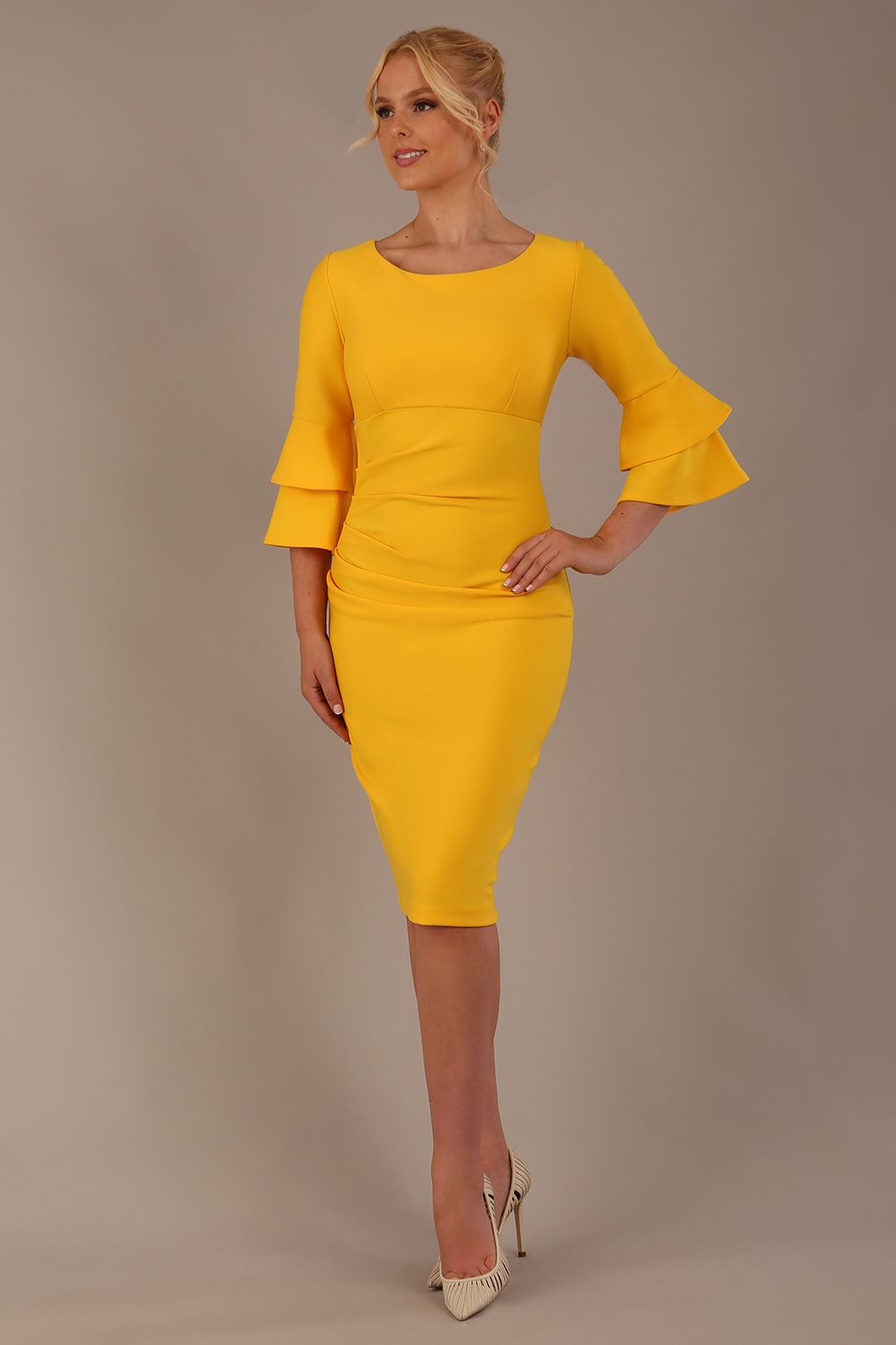 A woman wearing a sunshine yellow knee-length diva catwalk meridian pencil dress with a round neckline, double layered peplum three quarter length sleeves, and standing against a plain background.