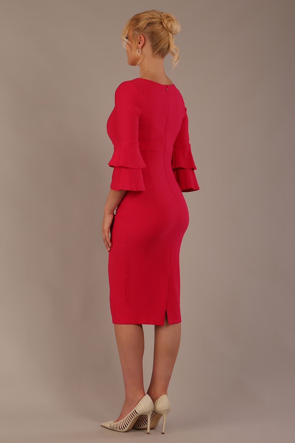 A woman wearing a yarrow pink knee-length diva catwalk meridian pencil dress with a round neckline, double layered peplum three quarter length sleeves, and standing against a plain background.
