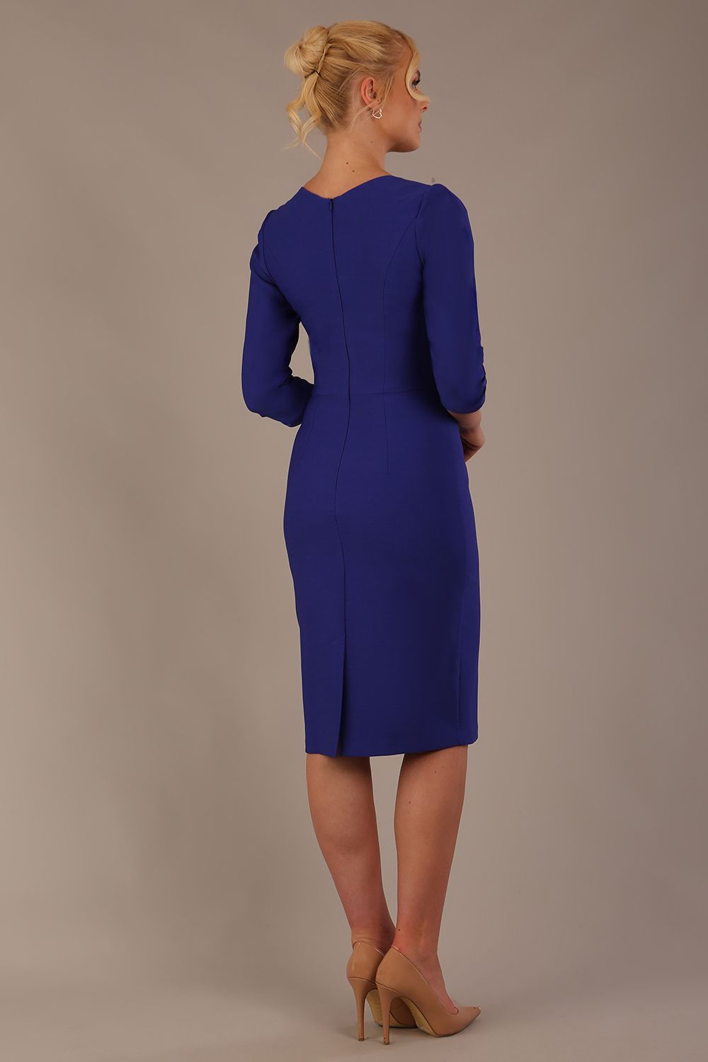A woman modeling a palace blue diva catwalk castleford pencil dress with an asymmetric neckline, bow waistband, and pleated skirt