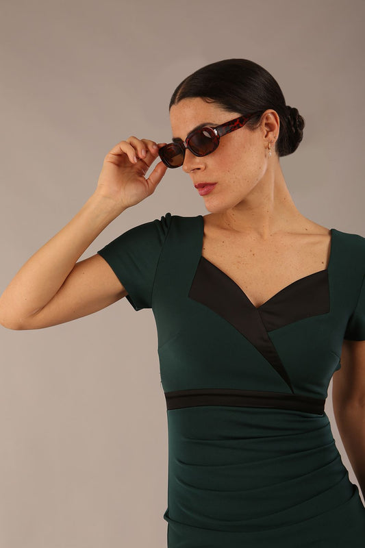 model is wearing a diva catwalk dress in forest green with black satin detail contrast on the neckline and waistband short sleeves and pleating details with knee-length