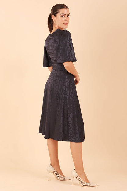 brunette model wearing diva catwalk cambrian satin swing dress with bell short sleeve wide asymmetric waistband and pleating under bust and low v-neckline in navy front