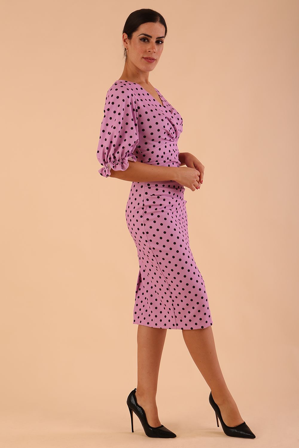 brunette model wearing diva catwalk palacio pencil fitted dress with three quarter puffed sleeve and pleating across the body with overlapping v-neckline in pink polka dot front