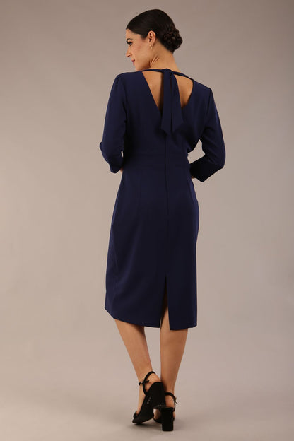 brunette model wearing diva catwalk wimpole a-line flattering dress with lowered v-neck and three quarter sleeve and cinching waistband and widened skirt with pleating in navy blue front