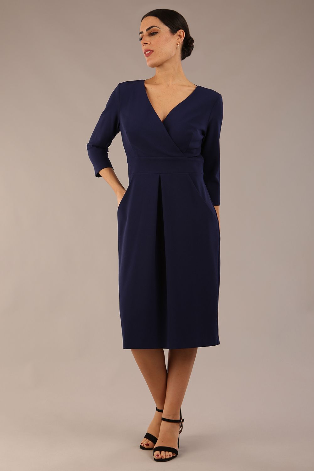 brunette model wearing diva catwalk wimpole a-line flattering dress with lowered v-neck and three quarter sleeve and cinching waistband and widened skirt with pleating in navy blue front