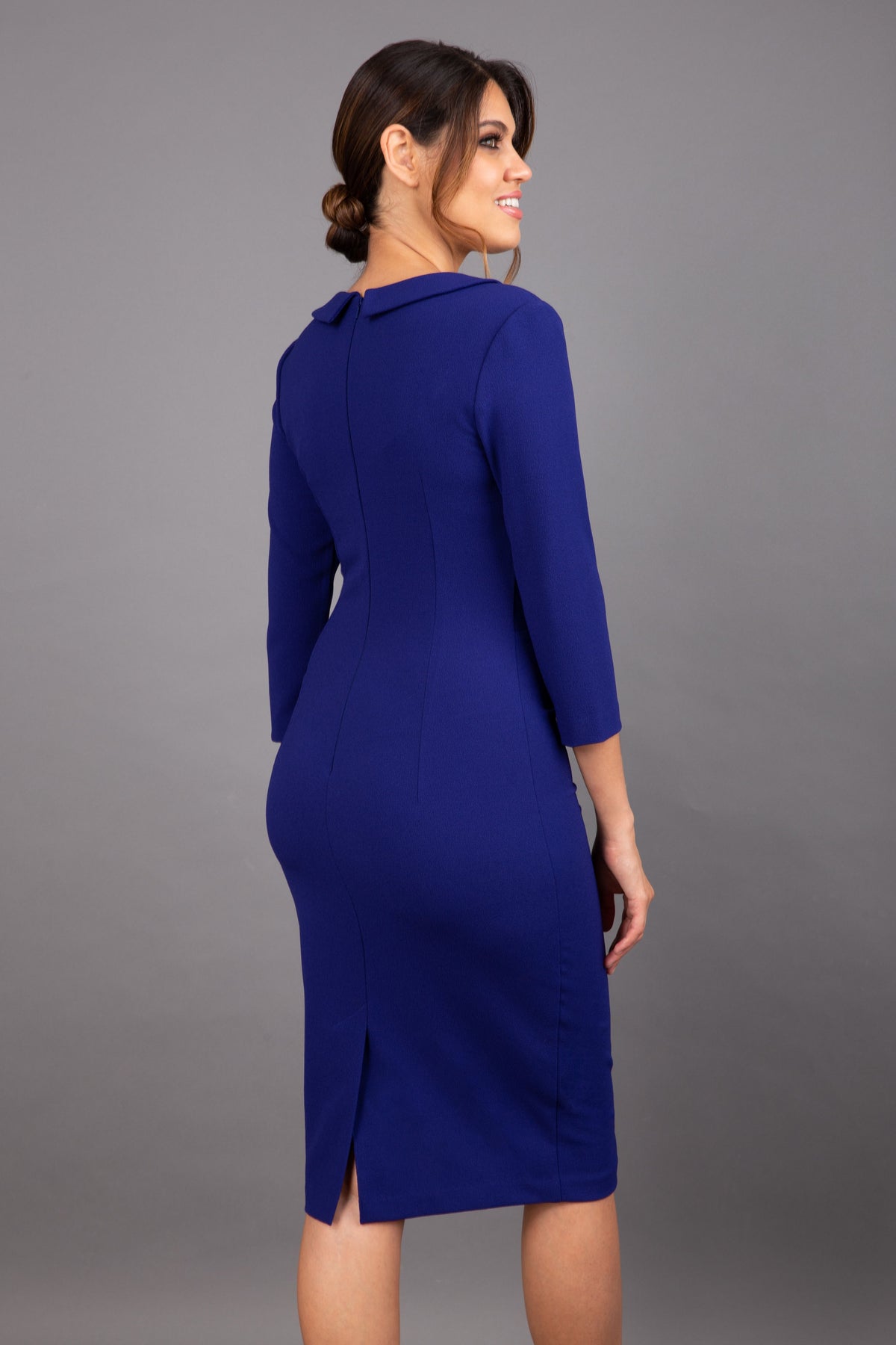 brunette model wearing diva catwalk elodie pencil fitted dress with rounded neckline and button detail and three quarter sleeve with pleating across the body from side triangle detail in oxford blue front
