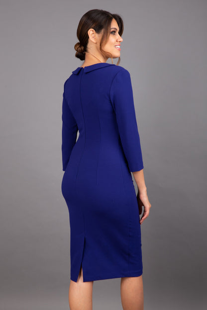 brunette model wearing diva catwalk elodie pencil fitted dress with rounded neckline and button detail and three quarter sleeve with pleating across the body from side triangle detail in oxford blue front