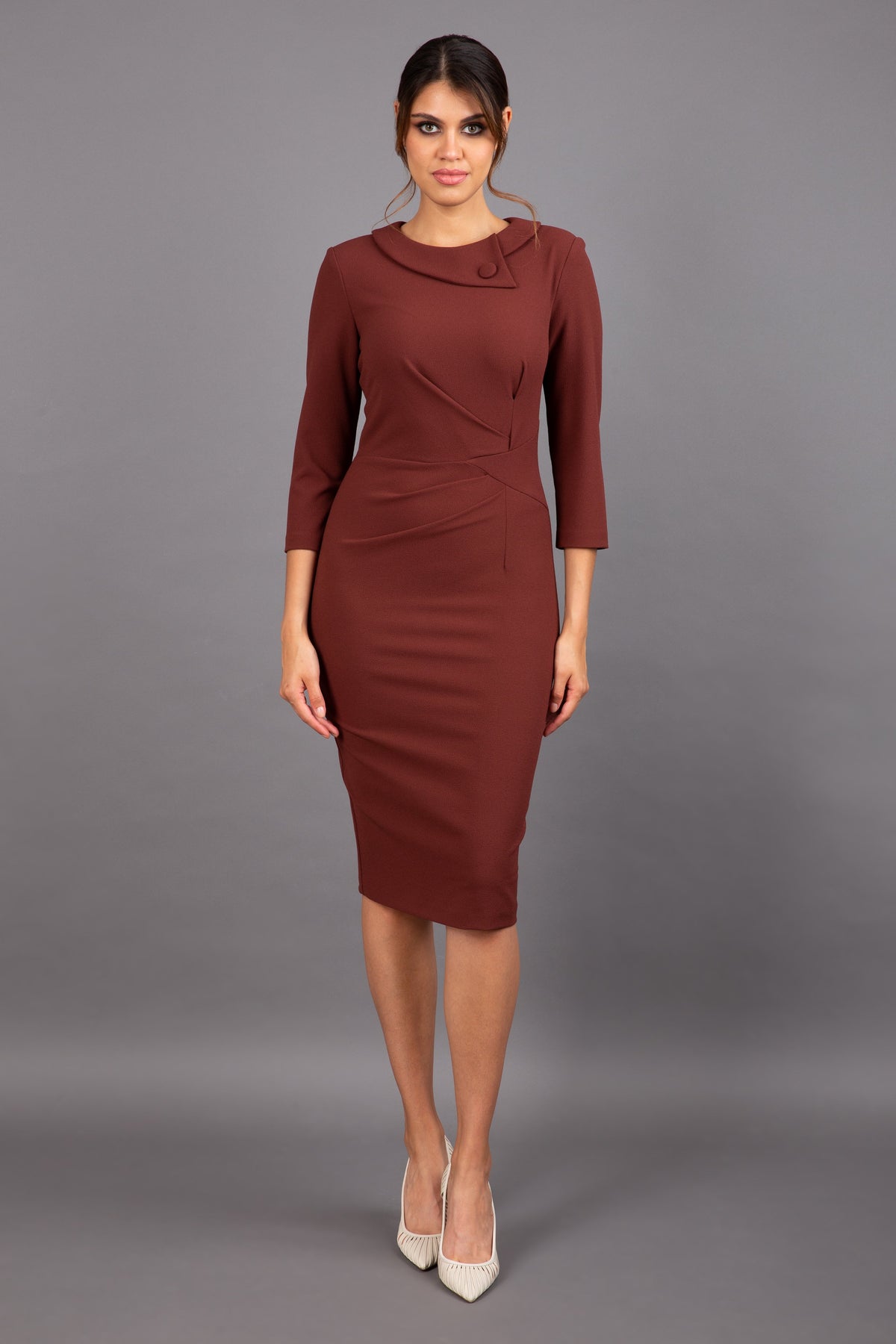 brunette model wearing diva catwalk elodie pencil fitted dress with rounded neckline and button detail and three quarter sleeve with pleating across the body from side triangle detail in mahogany front