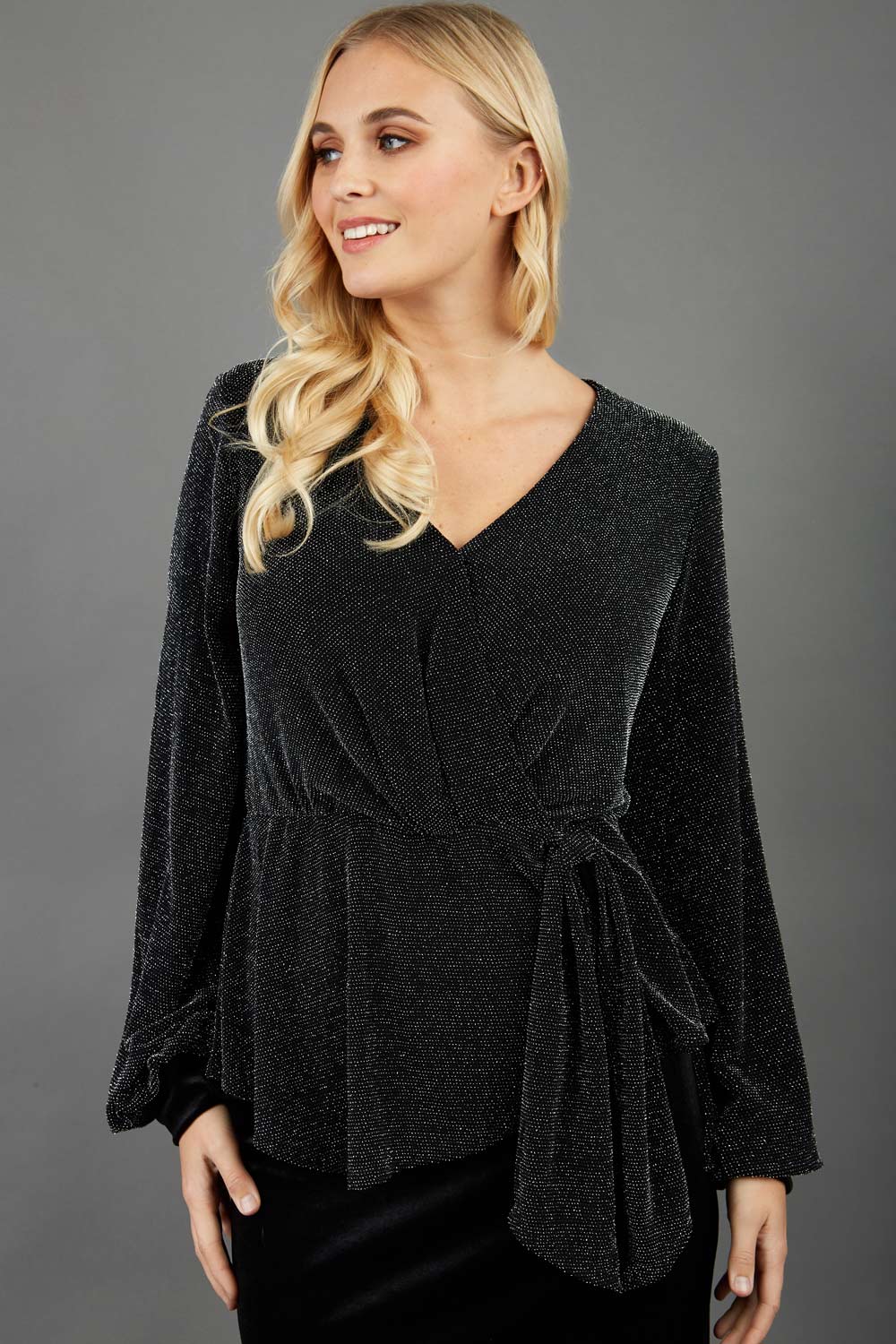 Blonde hair model is wearing a jersey sparkle v neck wrap blouse with bishop sleeve detail and bow on a side front image