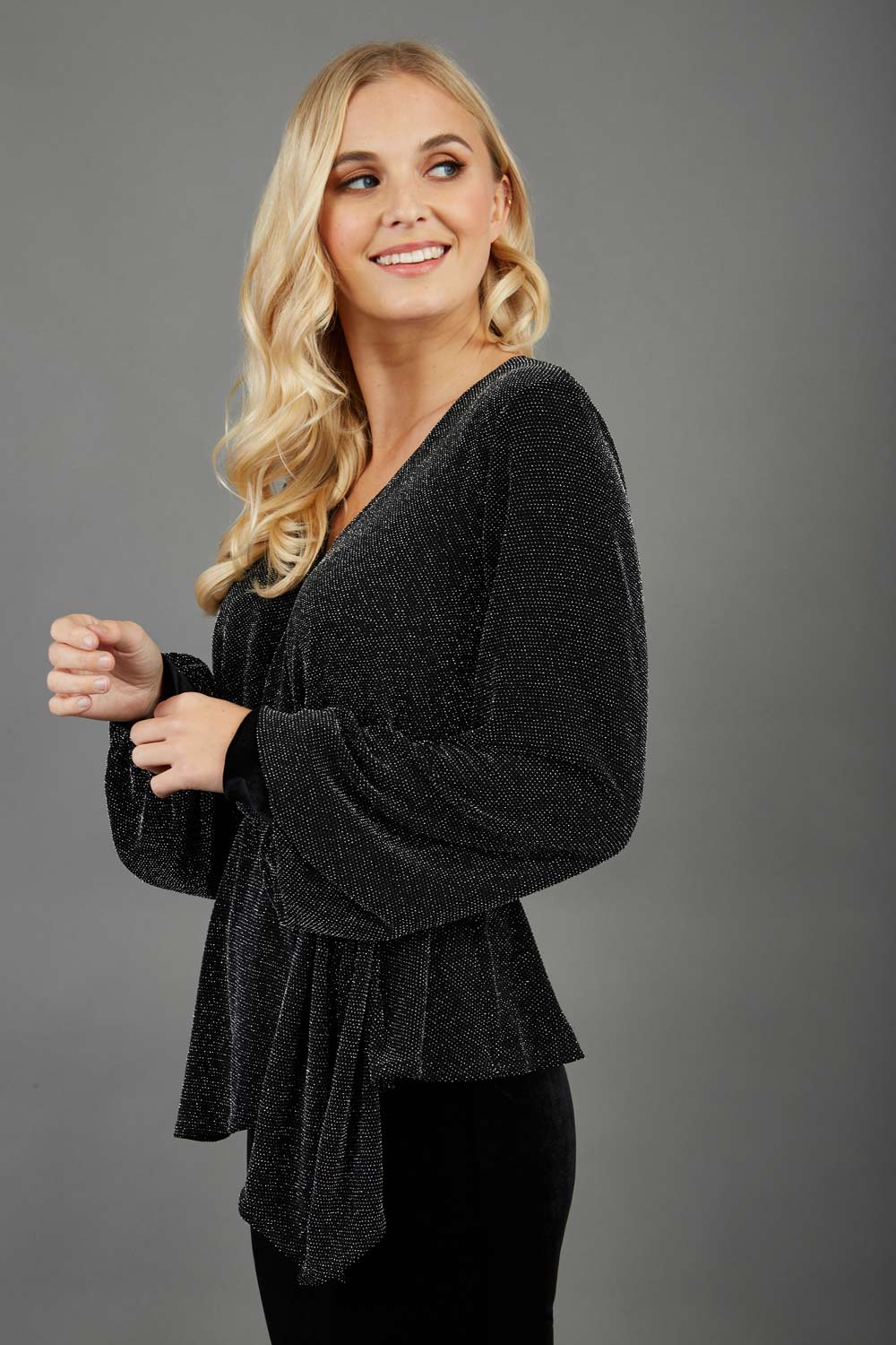 Blonde hair model is wearing a jersey sparkle v neck wrap blouse with bishop sleeve detail and bow on a side front image