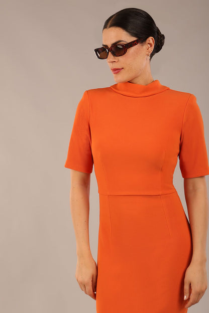 brunette model is wearing a diva catwalk Highmoor Short Sleeved Pencil Dress high neckline and shoulders pad in amber orange colour back image