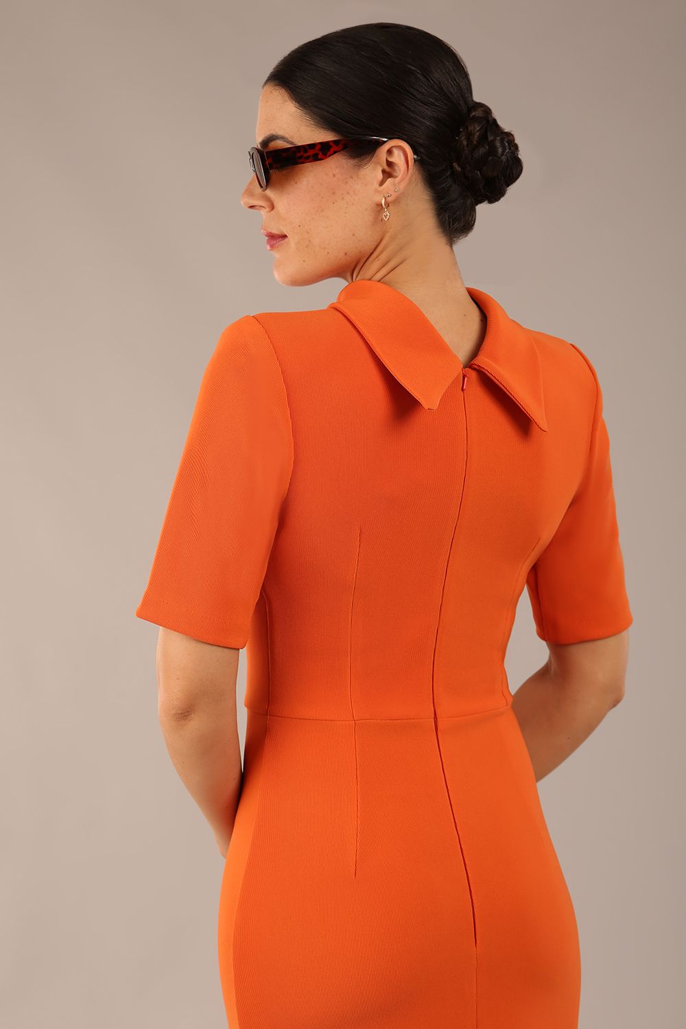brunette model is wearing a diva catwalk Highmoor Short Sleeved Pencil Dress high neckline and shoulders pad in amber orange colour back image