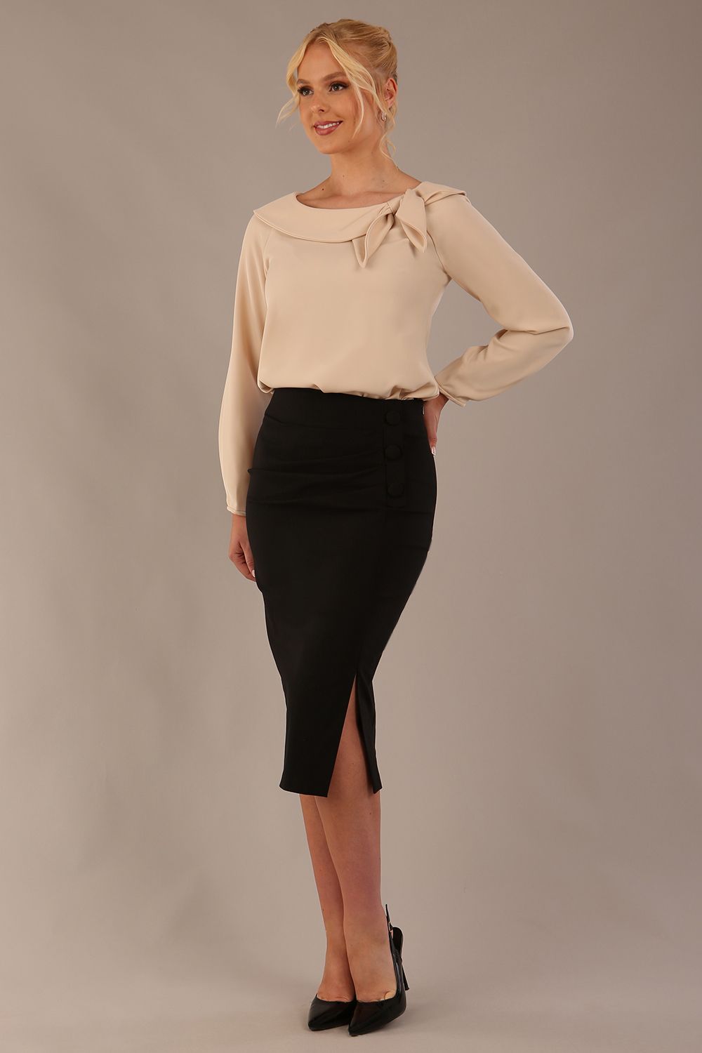 sitting model wearing a Cressida long sleeve top in almond cream, with a boat neckline and asymmetric bow, styled with a black skirt close up (Almond Cream)