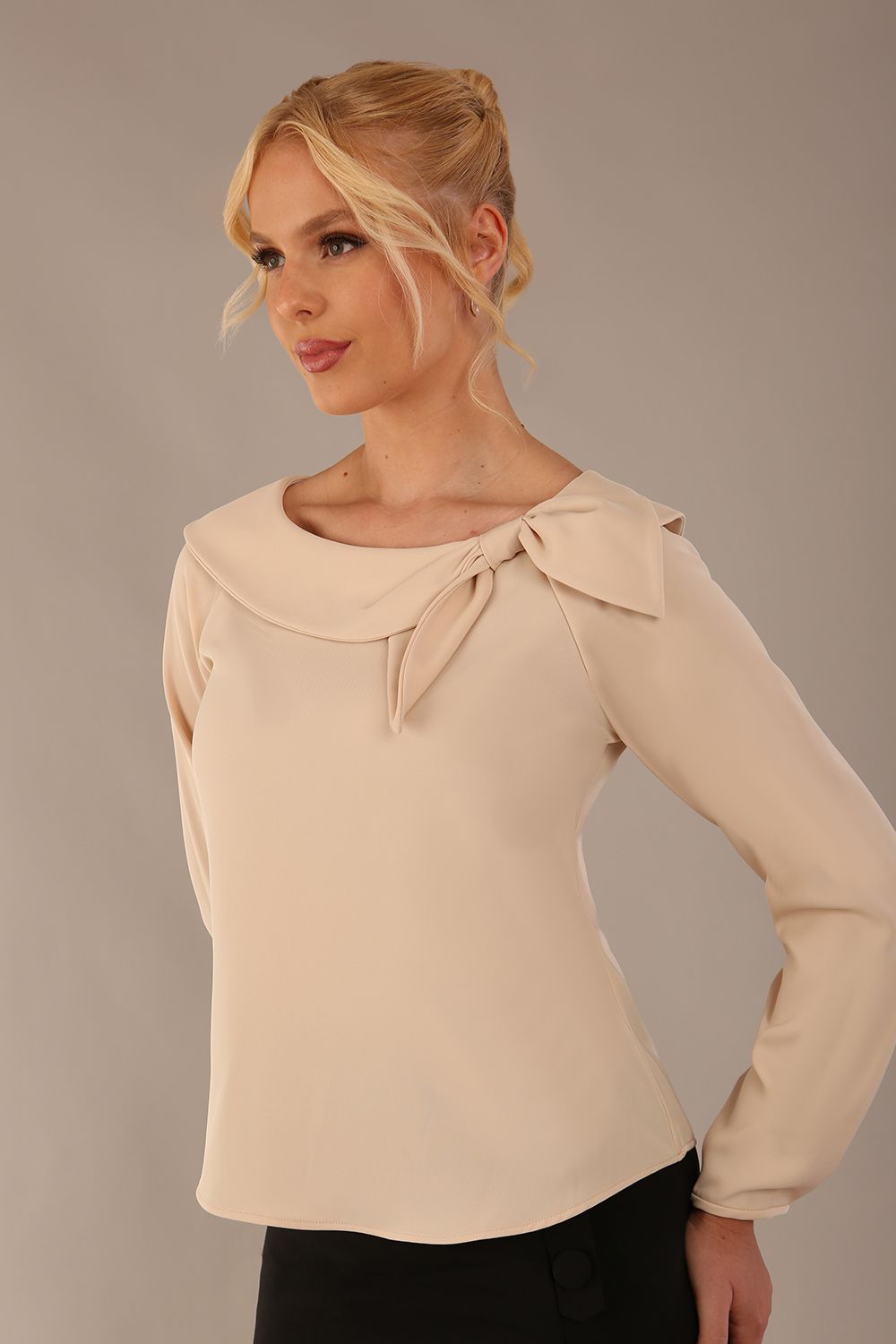 sitting model wearing a Cressida long sleeve top in almond cream, with a boat neckline and asymmetric bow, styled with a black skirt close up (Almond Cream)