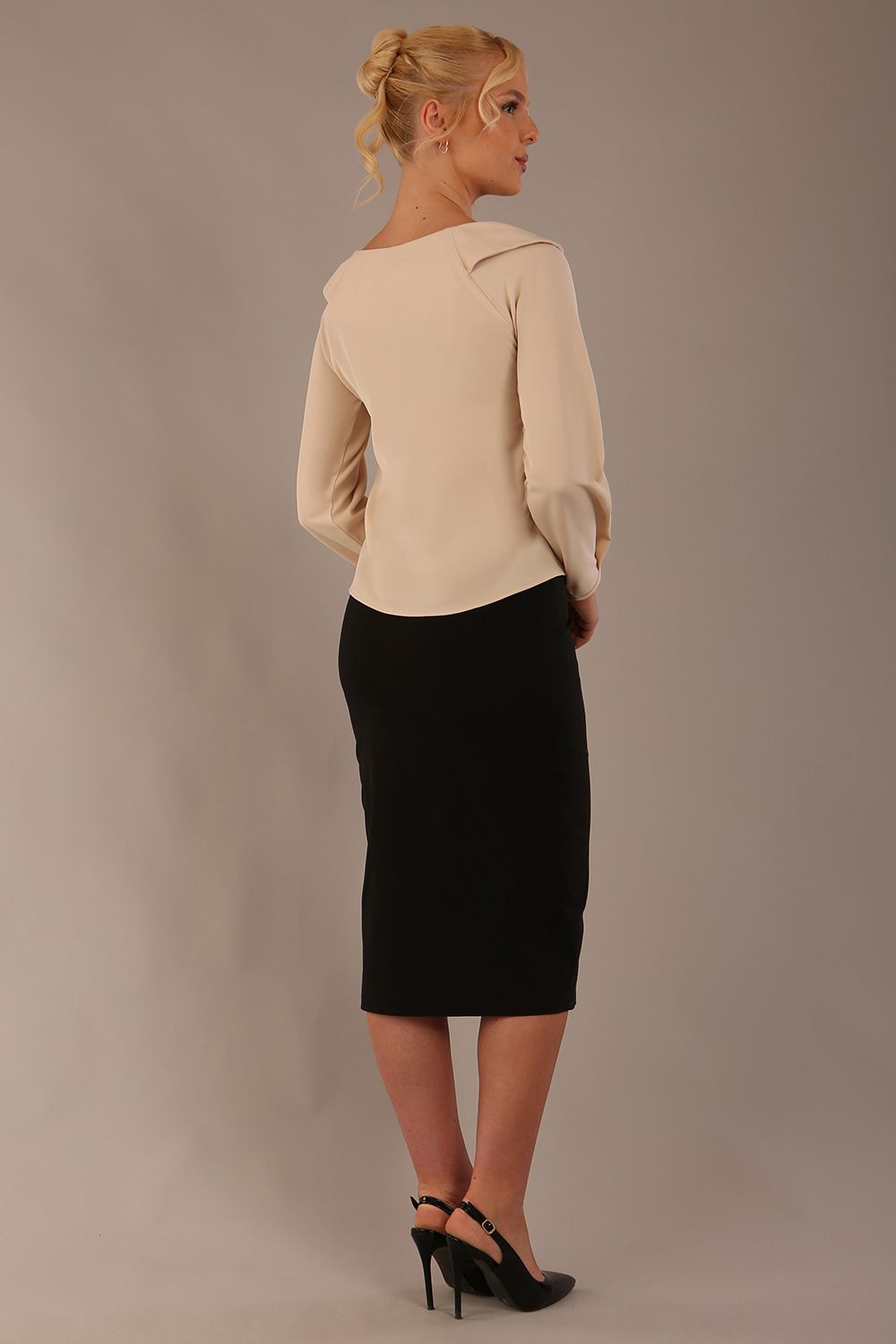 sitting model wearing a Cressida long sleeve top in almond cream, with a boat neckline and asymmetric bow, styled with a black skirt close up (Almond Cream)