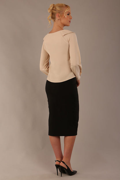 sitting model wearing a Cressida long sleeve top in almond cream, with a boat neckline and asymmetric bow, styled with a black skirt close up (Almond Cream)