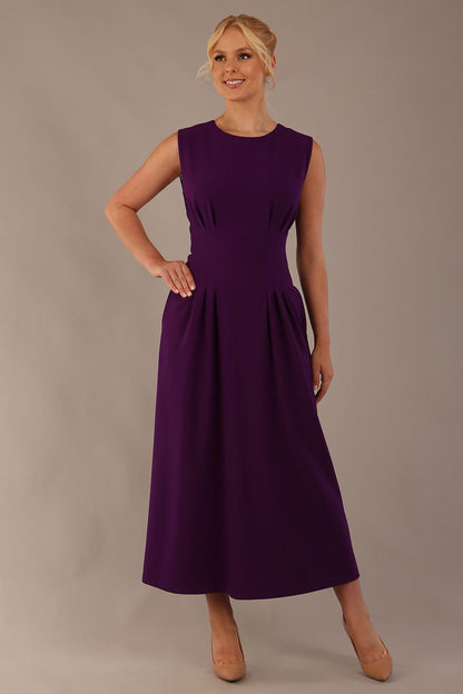 Model wearing the Cleo pleated midaxi dress in purple with hands in the discreet side pockets sitting on a chair against a plain background (Purple)