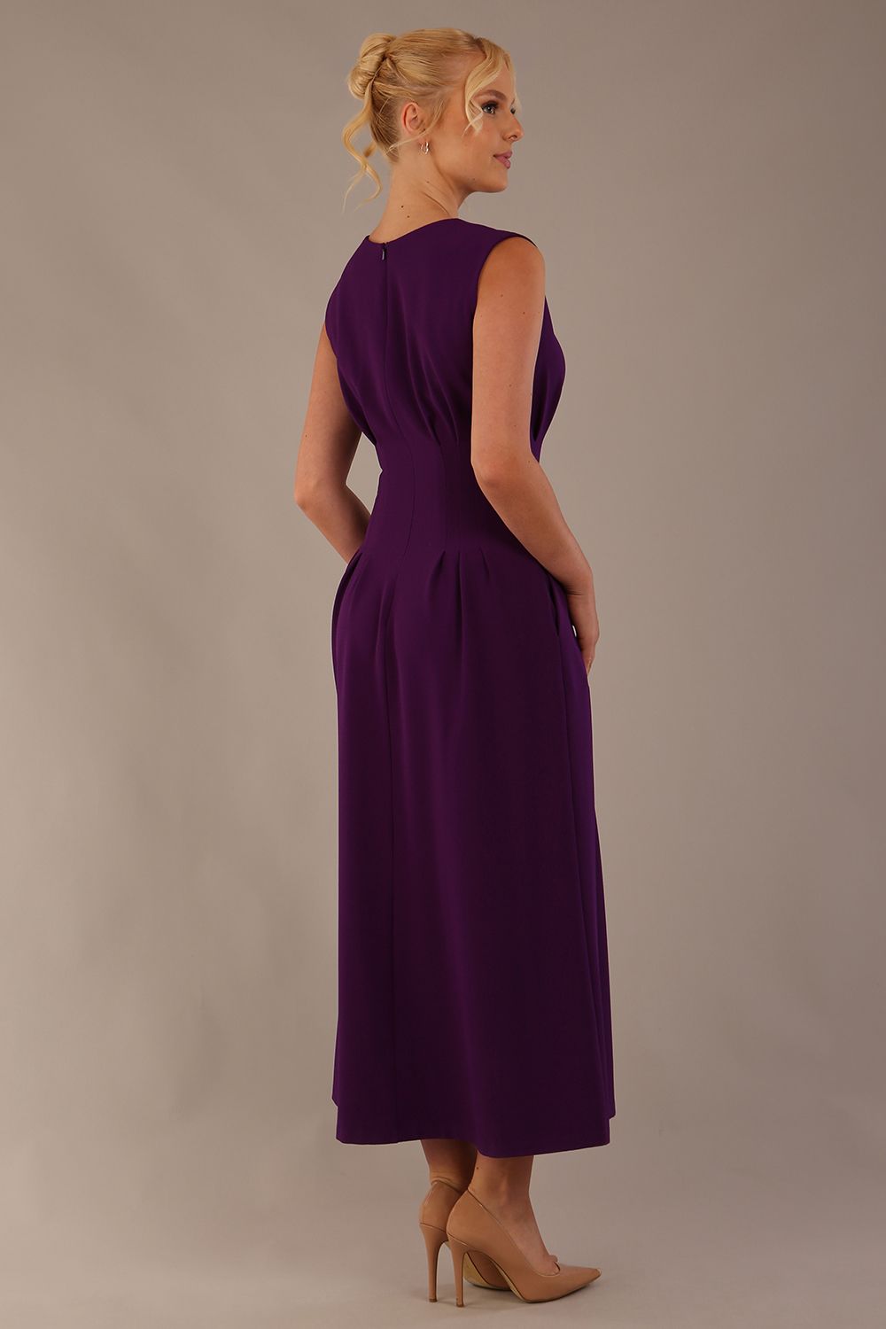 Model wearing the Cleo pleated midaxi dress in purple with hands in the discreet side pockets sitting on a chair against a plain background (Purple)