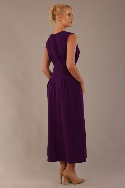 Model wearing the Cleo pleated midaxi dress in purple with hands in the discreet side pockets sitting on a chair against a plain background (Purple)