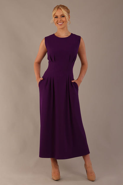 Model wearing the Cleo pleated midaxi dress in purple with hands in the discreet side pockets sitting on a chair against a plain background (Purple)