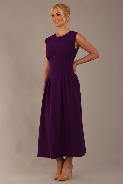 Model wearing the Cleo pleated midaxi dress in purple with hands in the discreet side pockets sitting on a chair against a plain background (Purple)