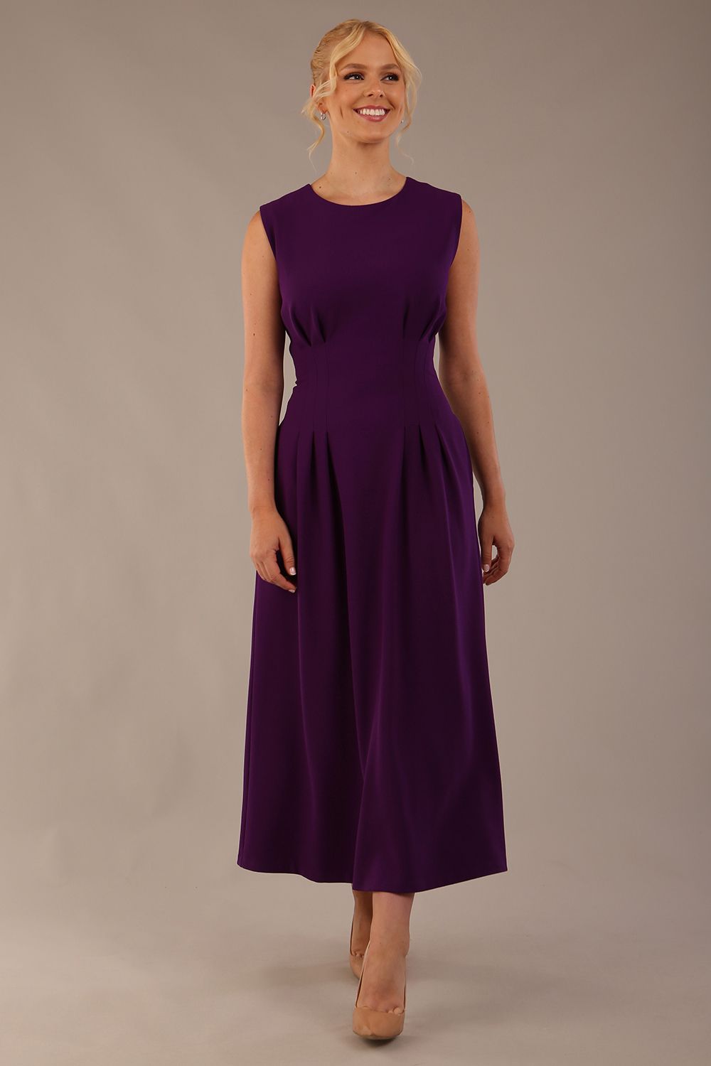 Model wearing the Cleo pleated midaxi dress in purple with hands in the discreet side pockets sitting on a chair against a plain background (Purple)