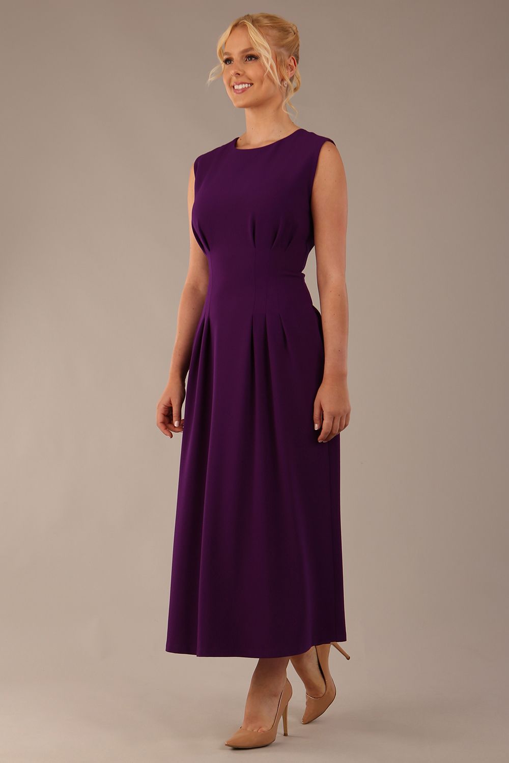 Model wearing the Cleo pleated midaxi dress in purple with hands in the discreet side pockets sitting on a chair against a plain background (Purple)
