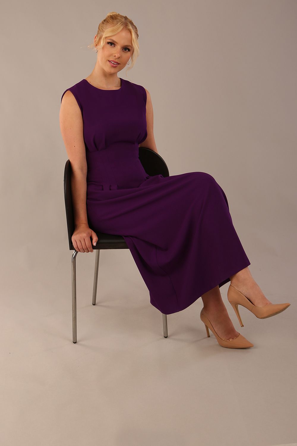 Model wearing the Cleo pleated midaxi dress in purple with hands in the discreet side pockets sitting on a chair against a plain background (Purple)
