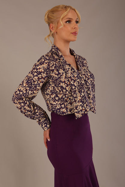Woman wearing a floral blouse with a plain background: The Truro top's unique draped V-neck with its two elegant front ties. Close-up view (Meadow Mischief Print)