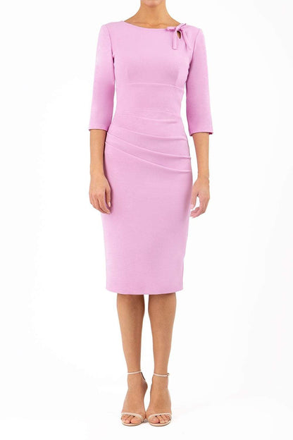 A model is wearing a three quarter sleeve pencil dress with a round neckline and bow detail at the front in pink