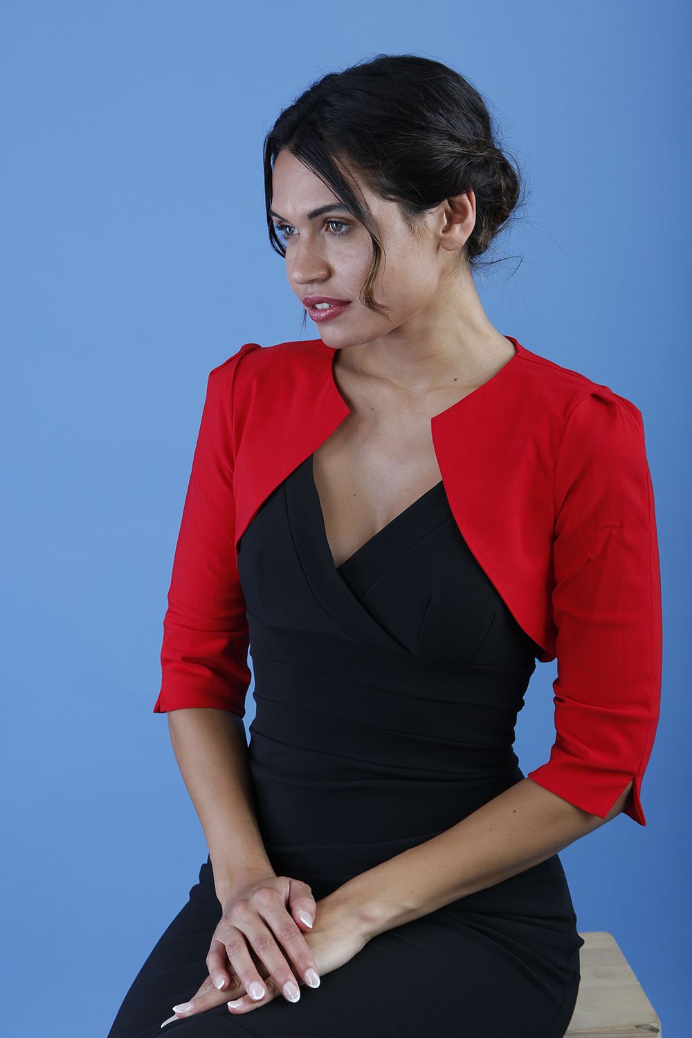 A woman wearing a red cropped bolero jacket with 3/4 sleeves and black clothing underneath. The photo is taken against a blue background.