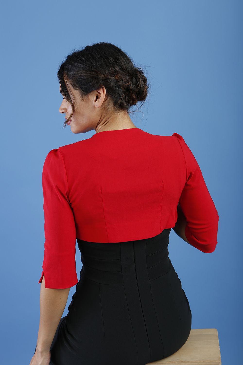 A woman wearing a red cropped bolero jacket with 3/4 sleeves and black clothing underneath. The photo is taken against a blue background.