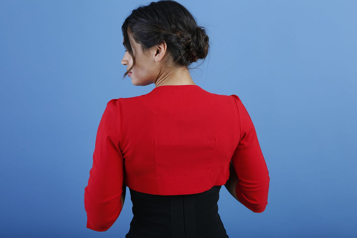 A woman wearing a red cropped bolero jacket with 3/4 sleeves and black clothing underneath. The photo is taken against a blue background.