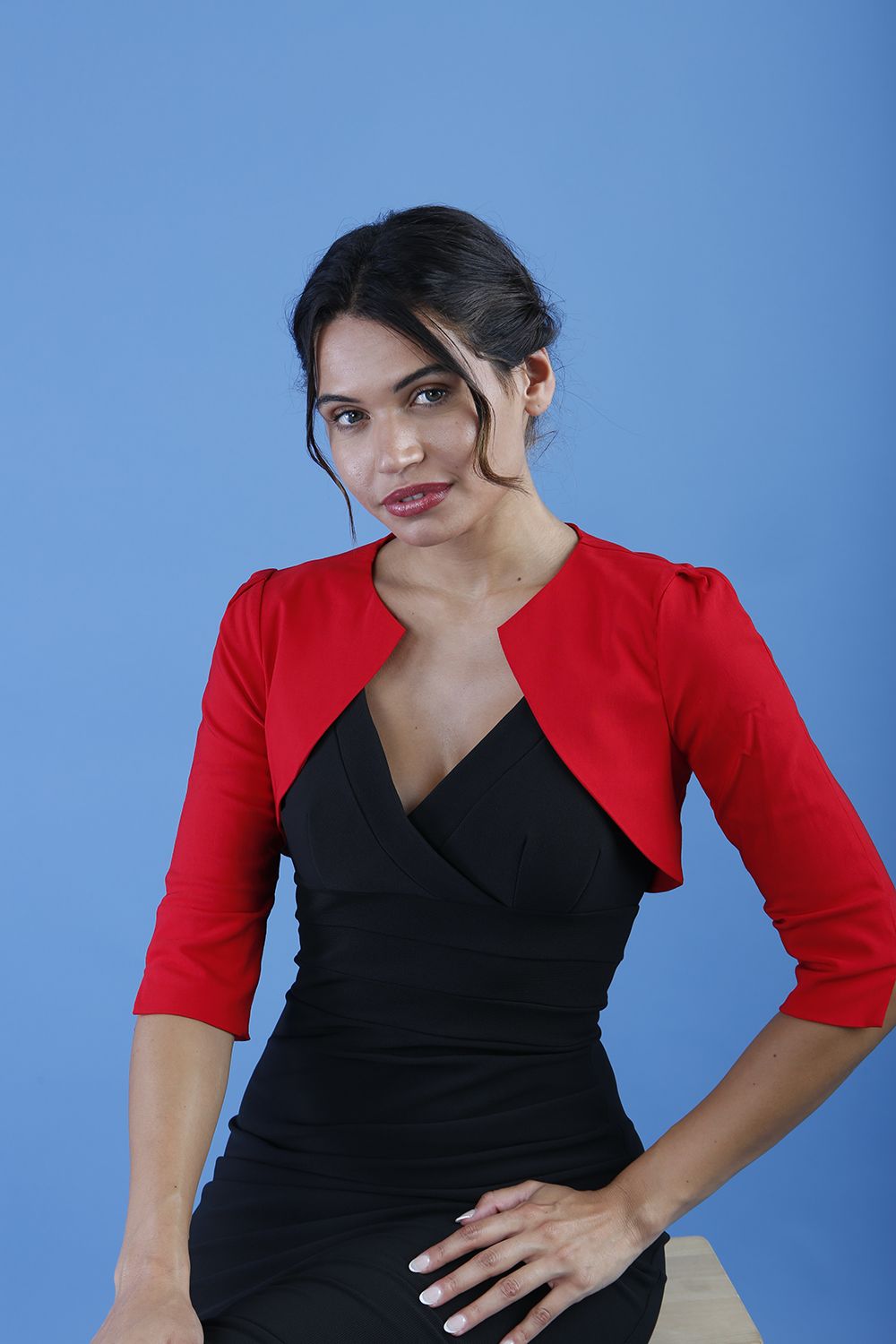 A woman wearing a red cropped bolero jacket with 3/4 sleeves and black clothing underneath. The photo is taken against a blue background.
